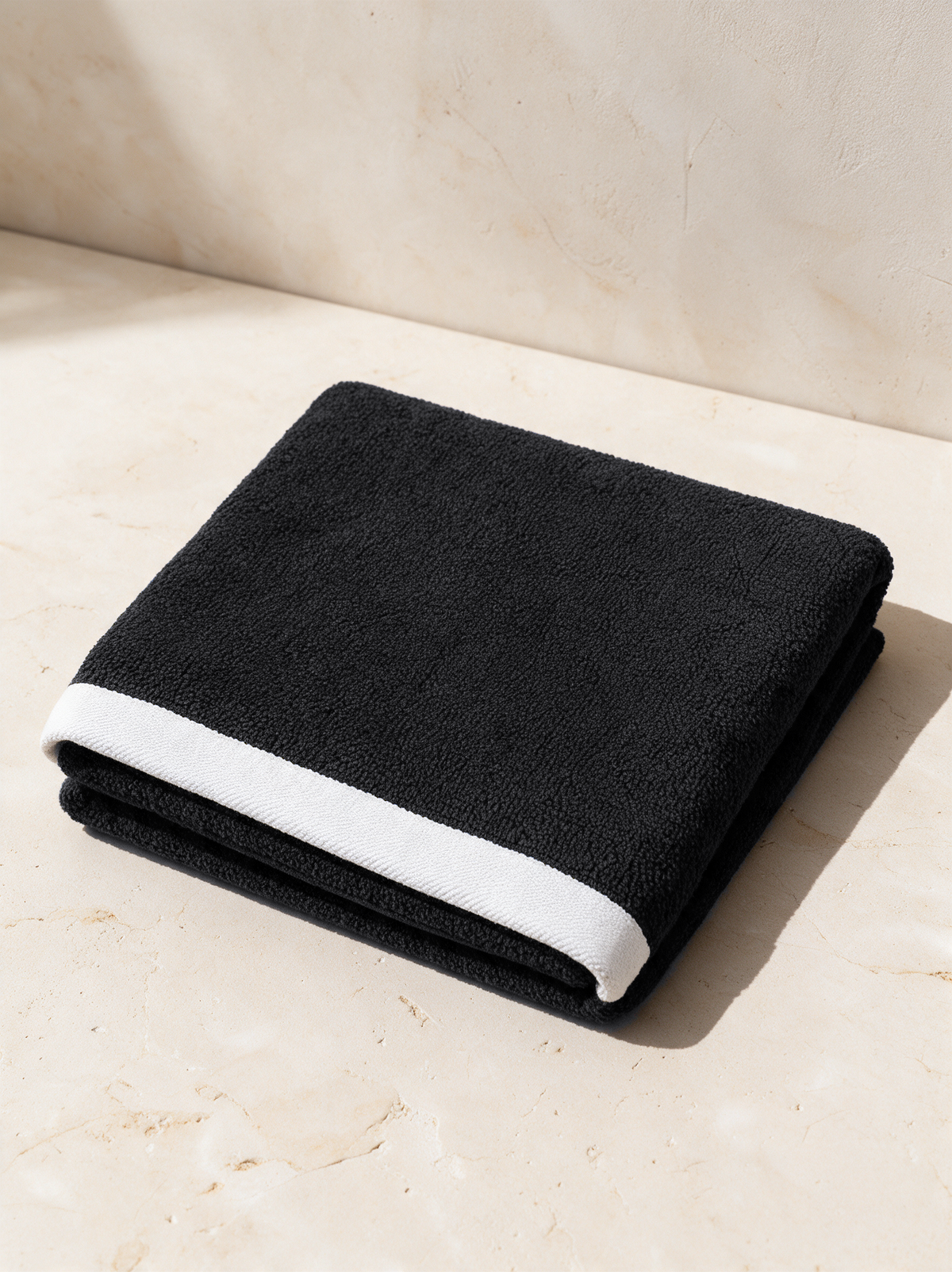 The Beach Towel in black with its white border, folded on warm stone