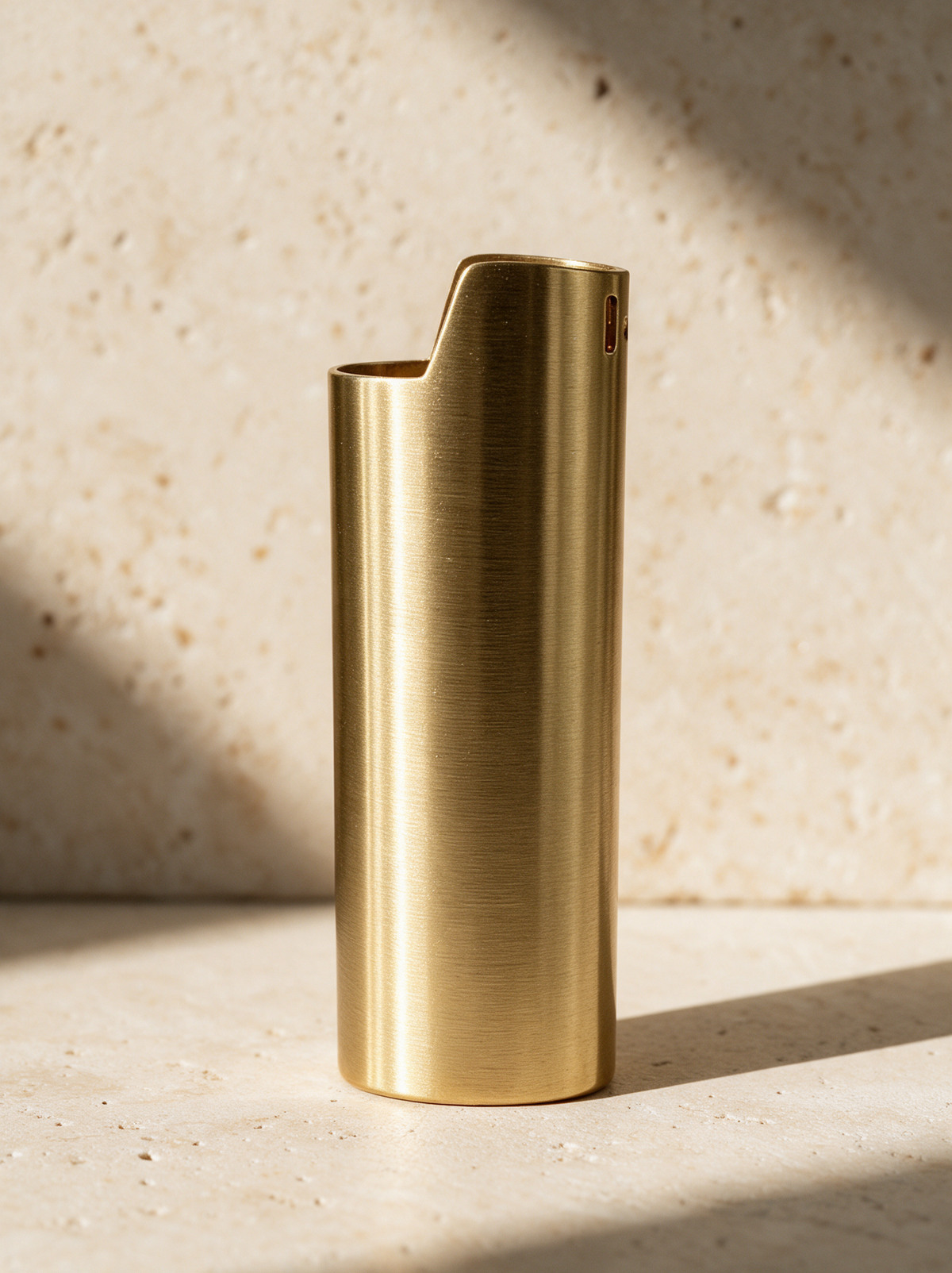 The Lighter Cover in brushed gold, standing on warm stone